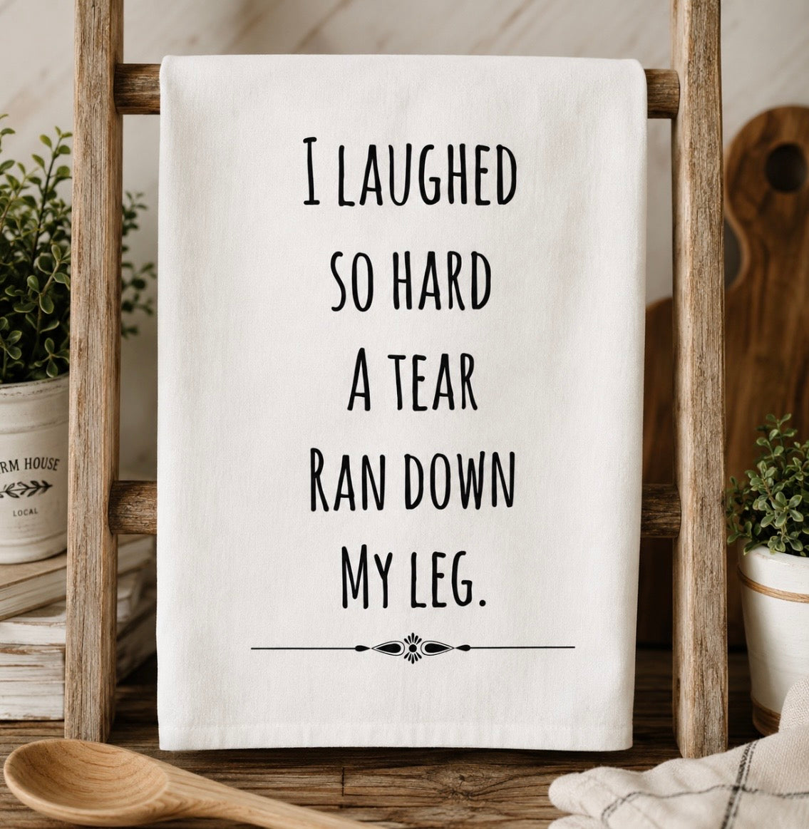 White kitchen towel with I Laughed So Hard A Tear Ran Down My Leg text displayed on a wooden ladder rack