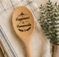 Engraved wooden spoon with Happiness is Homemade text and leaf detail on a linen background with greenery