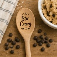 Engraved wooden spoon with Stir Crazy text and heart on a rustic wooden surface with chocolate chips
