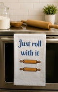 White kitchen towel with Just Roll With It text and rolling pin illustrations hanging on an oven door