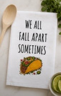 White kitchen towel with We All Fall Apart Sometimes text and taco illustration on a white surface with lime and herbs